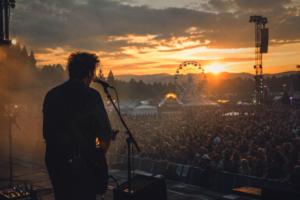 Musician performing on an outdoor festival stage at sunset, facing a large crowd with a Ferris wheel and mountains in the distance, representing live event and entertainment insurance.